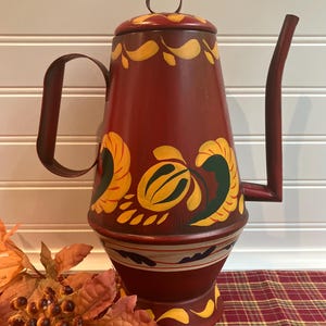 May include: A tall, red metal teapot with a long spout and a curved handle. The teapot is decorated with yellow and green floral designs. The lid has a small loop handle. The teapot sits on a red and gold plaid tablecloth.