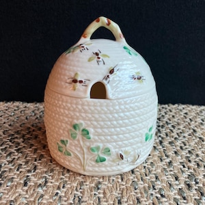 May include: A white ceramic honey pot shaped like a beehive with a brown handle and green shamrock designs. The pot is decorated with small brown bees.