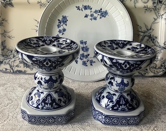 May include: A pair of ceramic candle holders featuring a white base with intricate dark blue floral and geometric patterns. Each holder has a wide, circular top, a rounded central stem, and a square base with detailed border designs.