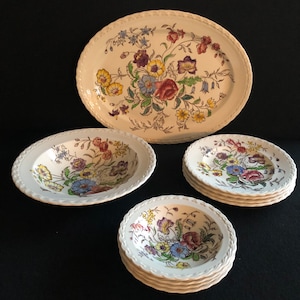 May include: A set of vintage floral patterned dinnerware. The set includes an oval serving platter, a large bowl, a stack of small bowls, and a stack of small plates. The floral pattern features red, yellow, blue, and purple flowers on a cream background.