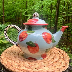 May include: A light blue ceramic teapot with a red rim and lid, decorated with red apple illustrations. The handle has a black and white zebra stripe pattern. The teapot is sitting on a woven placemat.