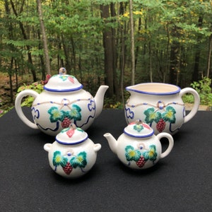 May include: A white ceramic tea set with a grapevine design. The set includes a teapot, creamer, and sugar bowl. The pieces are decorated with green leaves, blue vines, and red berries.