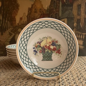 May include: White ceramic bowl with a green and orange lattice border. The bowl features a central image of a basket overflowing with pears, apples, grapes, and cherries. A stack of bowls is visible in the background.
