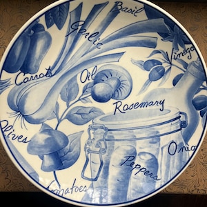 May include: White ceramic plate with a blue and white illustration of various cooking ingredients, including garlic, carrots, olives, and a jar. The plate has the names of the ingredients written in cursive script around the images.