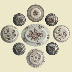 May include: A collection of vintage ceramic plates and serving dishes. The set includes various sizes and shapes, featuring floral and landscape designs in shades of red, brown, and cream. The oval platter is adorned with a large floral arrangement.