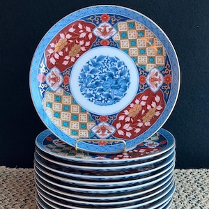 May include: A stack of ten white ceramic plates with a blue, red, and gold floral pattern. The plates have a black rim and are stacked on a woven surface.