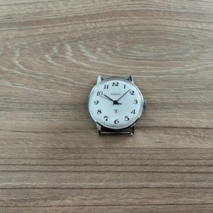 Puede incluir: Un reloj de pulsera plateado con una esfera blanca y números negros. El reloj tiene una correa plateada y una pequeña estrella en la posición de las 12 en punto.