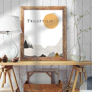 May include: A framed print with a minimalist design featuring a yellow sun, gray mountains, and the text "Friluftsliv". The print is hanging on a white wood wall above a wooden table with a lamp and a vase.