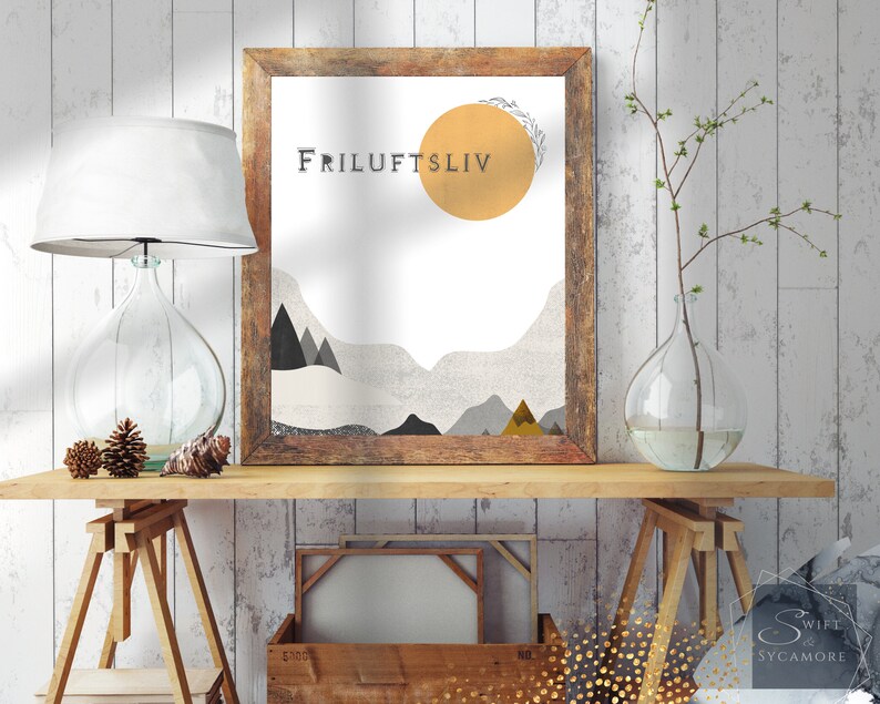 May include: A framed print with a minimalist design featuring a yellow sun, gray mountains, and the text "Friluftsliv". The print is hanging on a white wood wall above a wooden table with a lamp and a vase.