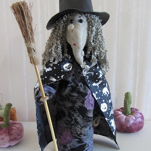 May include: A handmade Halloween witch doll with a black hat, grey curly hair, and a long nose. She wears a black and purple patterned dress and a cape with bats and stars. The witch holds a broom and is flanked by purple pumpkins.