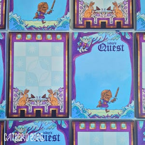 May include: A rectangular notepad with a purple border featuring a knight in gold armour holding a sword, a small green creature in a tree, and the text "today's QUEST". The design includes illustrations of rabbits and floral elements in a fantasy style.