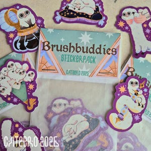 Puede incluir: Una colección de pegatinas caprichosas con criaturas de estilo caricaturesco con ojos grandes. Las pegatinas son de varias formas y tamaños, con una paleta de colores predominante de blanco, morado y dorado. También se ve un paquete de pegatinas con el texto "Brushbuddies STICKER PACK".
