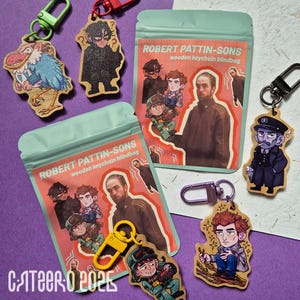 Könnte beinhalten: Eine Sammlung von Holzschlüsselanhängern und Blind Bags. Die Schlüsselanhänger zeigen verschiedene Cartoon-Illustrationen und sind an Metallverschlüssen befestigt. Die Blind Bags sind hellgrün mit dem Text "ROBERT PATTIN-SONS wooden keychain blindbag".