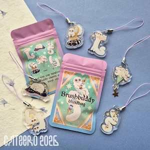 Può includere: Diversi ciondoli in acrilico trasparente con vari disegni di personaggi dei cartoni animati, ciascuno attaccato a un cordino lavanda. Sono visibili anche due sacchetti rosa con la scritta "Brushbuddy blindbag". Lo sfondo è blu chiaro.