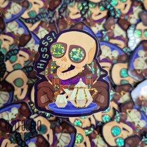 Peut inclure: Autocollant représentant un crâne de dessin animé avec des lunettes de soleil scintillantes et le mot "HISSS!". Le crâne est assis et porte une écharpe violette. Une théière et une tasse sont sur un plateau bleu. Le sticker est marron et doré.