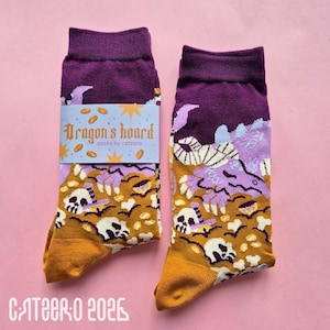 Könnte beinhalten: Ein Paar Socken mit lila Bündchen und Zehen, mit einem Drachen- und Totenkopf-Design. Die Socken tragen den Text "Dragon's hoard" auf einem Etikett. Der Hintergrund ist hellrosa.