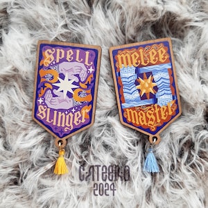Può includere: Due ornamenti in legno a forma di scudo con un design di lanciatore di incantesimi viola e arancione e un design di maestro di mischia blu e arancio. Ogni ornamento ha un fiocco che pende dalla parte inferiore.