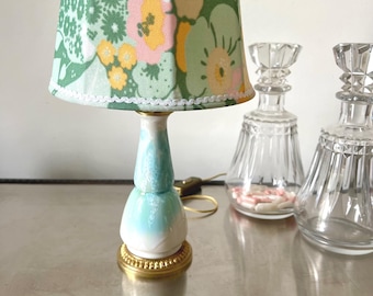Puede incluir: Una pequeña lámpara decorativa con una pantalla con estampado floral en tonos verdes, rosas y amarillos. La base es blanca y azul claro, con una base dorada. Dos botellas decorativas de vidrio transparente están en el fondo.