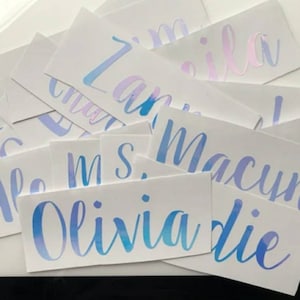 May include: A set of white stickers with blue and purple ombre lettering. The stickers feature the names Olivia, Die, Macyn, MS, and  Zara.