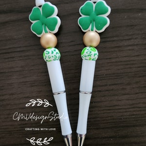 May include: Two white pens with green four-leaf clover charms, gold beads, and white beads with green spots. The pens have silver accents. The text "CM Design Studio Crafting with Love" is visible below the pens.