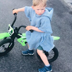 Pode incluir: Um menino pequeno vestindo um poncho azul claro com capuz monta uma bicicleta de terra verde e preta com rodas pretas. Ele est&aacute; usando sand&aacute;lias azuis.