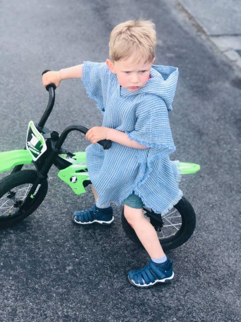 Pode incluir: Um menino pequeno vestindo um poncho azul claro com capuz monta uma bicicleta de terra verde e preta com rodas pretas. Ele est&aacute; usando sand&aacute;lias azuis.