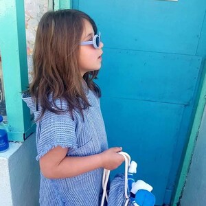 Pode incluir: Uma pessoa jovem usando uma toalha com capuz listrada de azul e branco, &oacute;culos de sol azuis e segurando uma toalha listrada de azul e branco. A pessoa est&aacute; em p&eacute; em frente a uma porta turquesa com uma placa vermelha e branca que diz "ICE CREAM".