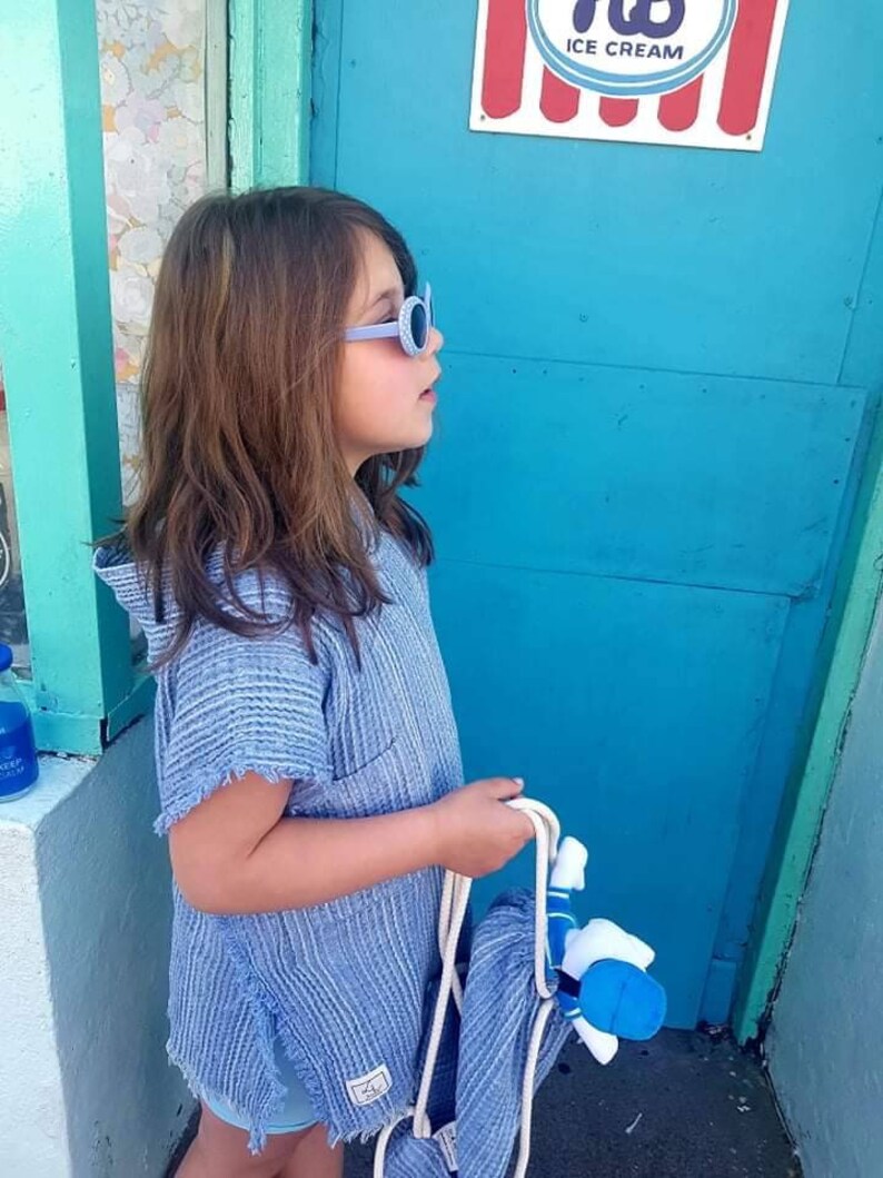 Pode incluir: Uma pessoa jovem usando uma toalha com capuz listrada de azul e branco, &oacute;culos de sol azuis e segurando uma toalha listrada de azul e branco. A pessoa est&aacute; em p&eacute; em frente a uma porta turquesa com uma placa vermelha e branca que diz "ICE CREAM".