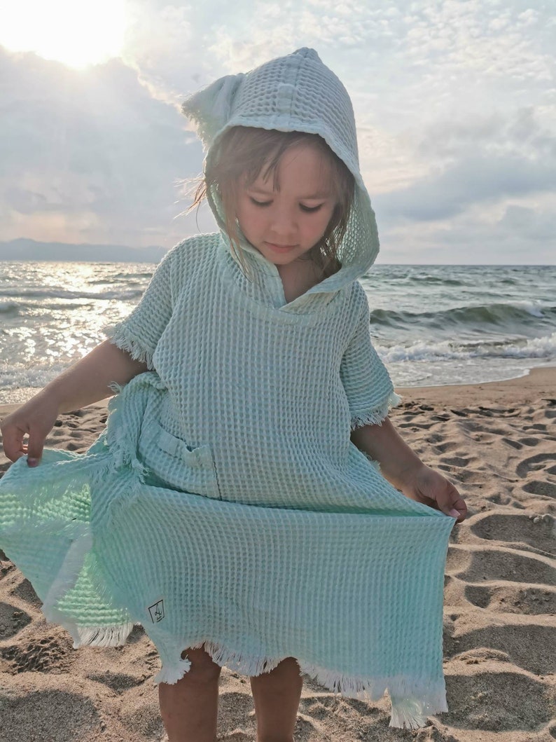 Pode incluir: Uma toalha com capuz azul claro com uma borda desfiada. A toalha &eacute; feita de tecido waffle e tem um pequeno bolso na frente. A toalha est&aacute; sendo usada por uma crian&ccedil;a em uma praia.