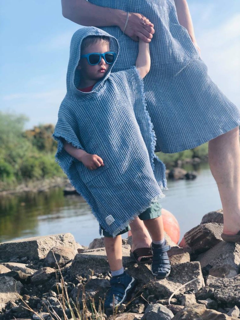 Pode incluir: Uma crian&ccedil;a vestindo uma toalha com capuz azul com uma borda desfiada, de m&atilde;os dadas com um adulto vestindo uma toalha semelhante. A crian&ccedil;a est&aacute; usando &oacute;culos de sol azuis e sapatos azuis e pretos. Eles est&atilde;o em p&eacute; em rochas perto de um corpo de &aacute;gua.