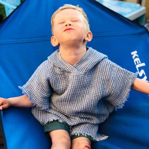 Pode incluir: Uma crian&ccedil;a pequena vestindo uma toalha com capuz listrada azul e branca est&aacute; sentada em uma cadeira de camping azul. A cadeira tem a palavra "Kelsey" impressa na lateral.