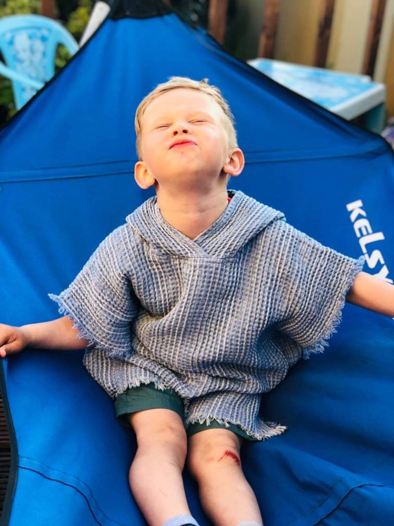 Pode incluir: Uma crian&ccedil;a pequena vestindo uma toalha com capuz listrada azul e branca est&aacute; sentada em uma cadeira de camping azul. A cadeira tem a palavra "Kelsey" impressa na lateral.