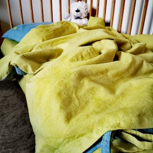 May include: A yellow linen crib sheet with a blue trim and a white stuffed bear sitting on top of it.
