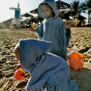 Pode incluir: Duas crian&ccedil;as vestindo toalhas com capuz azul brincam em uma praia de areia. Uma crian&ccedil;a est&aacute; segurando um brinquedo amarelo e laranja.