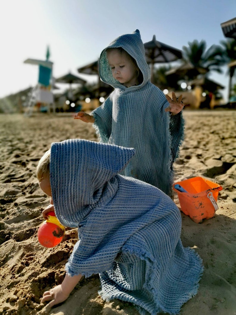 Pode incluir: Duas crian&ccedil;as vestindo toalhas com capuz azul brincam em uma praia de areia. Uma crian&ccedil;a est&aacute; segurando um brinquedo amarelo e laranja.