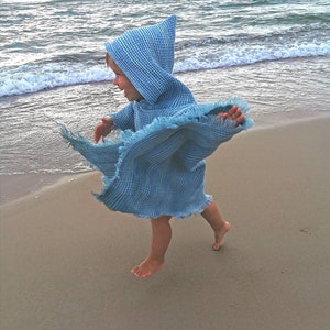 Pode incluir: Uma crian&ccedil;a usando uma toalha com capuz azul claro corre em uma praia de areia. A toalha tem uma borda franjada e &eacute; feita de tecido waffle.
