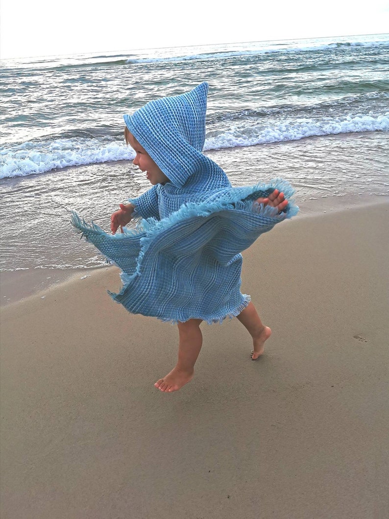 Pode incluir: Uma crian&ccedil;a usando uma toalha com capuz azul claro corre em uma praia de areia. A toalha tem uma borda franjada e &eacute; feita de tecido waffle.