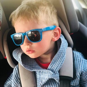 Pode incluir: Uma crian&ccedil;a pequena usando &oacute;culos de sol azuis e um su&eacute;ter com capuz azul est&aacute; sentada em uma cadeirinha de carro.