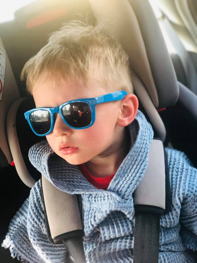Pode incluir: Uma crian&ccedil;a pequena usando &oacute;culos de sol azuis e um su&eacute;ter com capuz azul est&aacute; sentada em uma cadeirinha de carro.