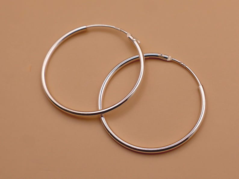 May include: A pair of polished silver hoop earrings with a circular design. The earrings feature a thin, smooth metallic band that forms a complete circle, with a small hinged clasp mechanism visible at the top of each hoop for secure closure.