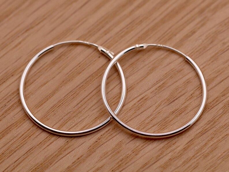 May include: A pair of thin, circular silver hoop earrings. Each earring features a smooth, polished metallic finish and a simple, continuous wire design with a small, integrated hinge mechanism for opening and closing the hoop securely.