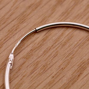 May include: A thin, circular silver hoop earring featuring a smooth, polished metallic finish. The earring includes a hinged closure mechanism with a small, visible hallmark stamp near the joint, showing the construction of the hoop.