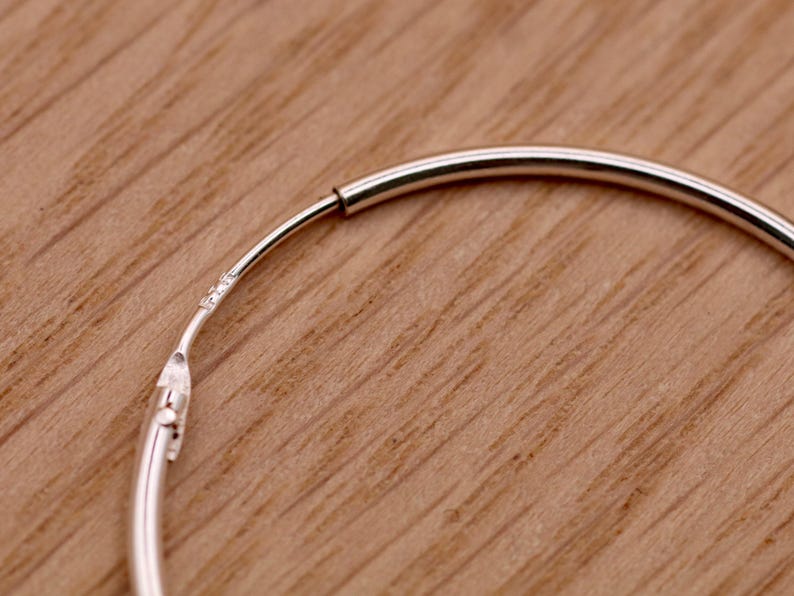 May include: A thin, circular silver hoop earring featuring a smooth, polished metallic finish. The earring includes a hinged closure mechanism with a small, visible hallmark stamp near the joint, showing the construction of the hoop.