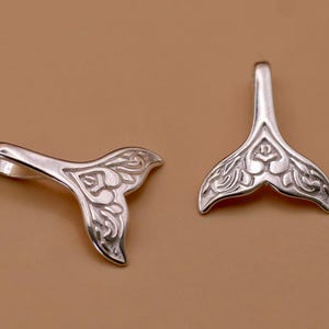 May include: Two silver whale tail pendants. Each pendant has a loop for a chain and an ornate floral design. The pendants are displayed on a light brown background. The pendants are likely for jewellery.