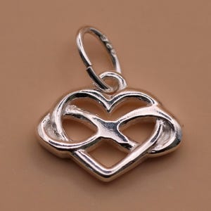 May include: A silver pendant featuring a heart and infinity symbol design. The pendant has a loop for a chain and is set against a neutral background. The pendant is made of a shiny metal.