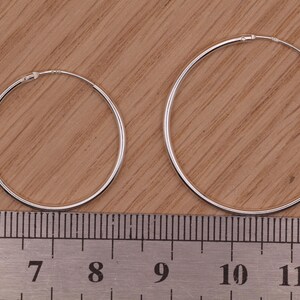 May include: Two thin silver hoop earrings of different sizes positioned above a metal ruler. The smaller hoop measures approximately 1.5 inches in diameter, while the larger hoop measures approximately 2.25 inches in diameter.