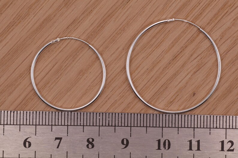 May include: Two thin silver hoop earrings of different sizes positioned above a metal ruler. The smaller hoop measures approximately 1.5 inches in diameter, while the larger hoop measures approximately 2.25 inches in diameter.