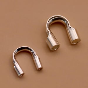 May include: Two silver-coloured, U-shaped jewellery findings. Each finding has a curved top and two open-ended cylindrical tubes at the bottom. The findings are displayed against a solid, light brown background.