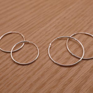 May include: Four thin, circular silver hoop earrings arranged in two pairs of different sizes. Each earring features a smooth, polished metallic finish and a simple, continuous wire design with a small, subtle closure mechanism visible on the side.
