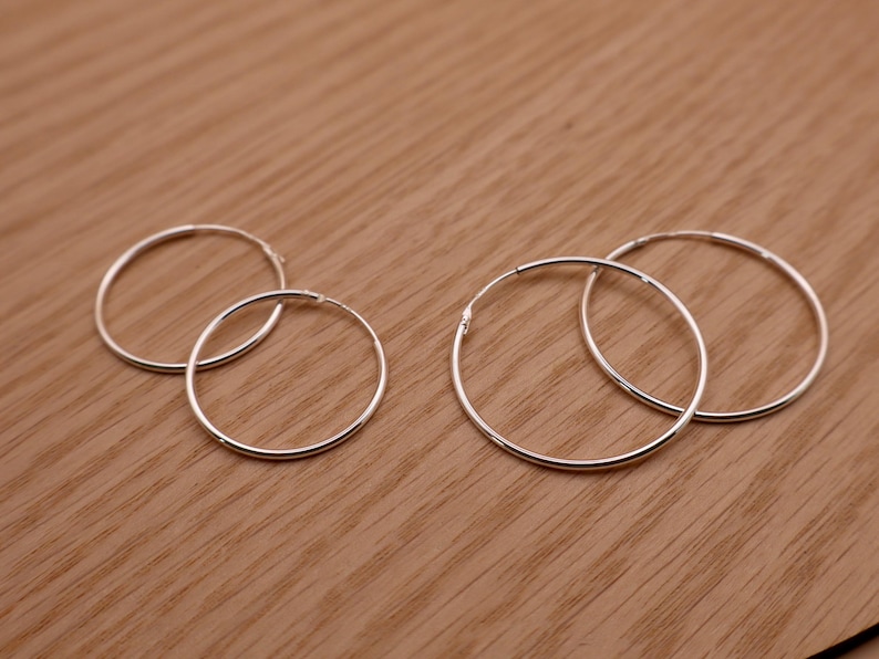 May include: Four thin, circular silver hoop earrings arranged in two pairs of different sizes. Each earring features a smooth, polished metallic finish and a simple, continuous wire design with a small, subtle closure mechanism visible on the side.